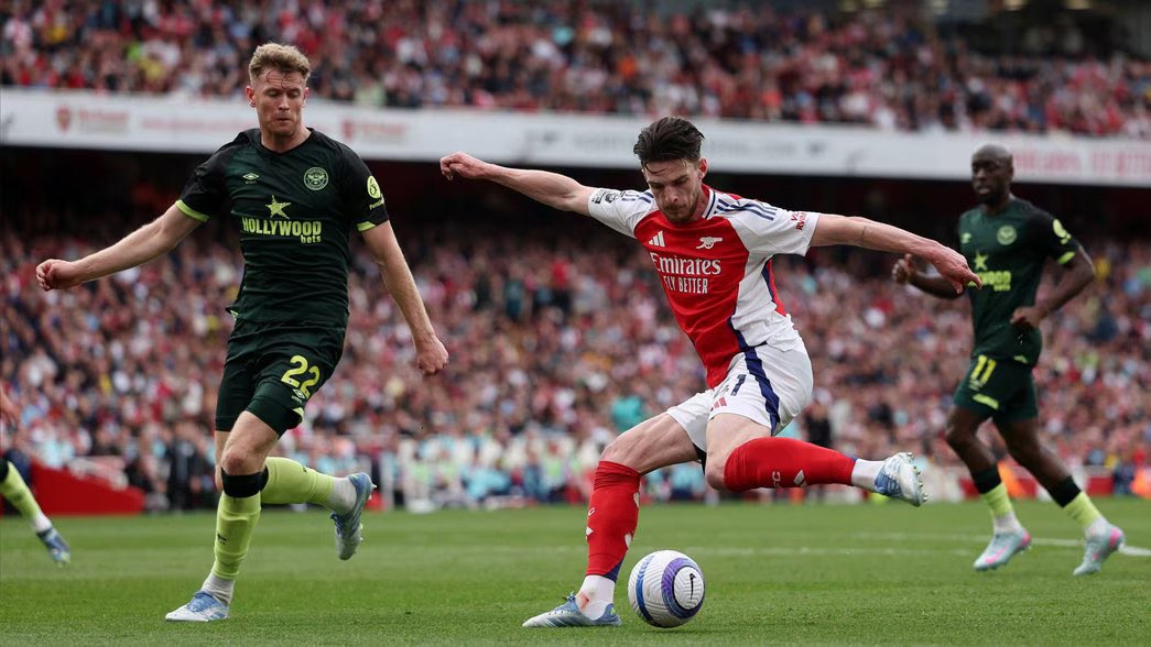 Arsenal midfielder Declan Rice kicking a ball in a stadium with Brentford player chasing